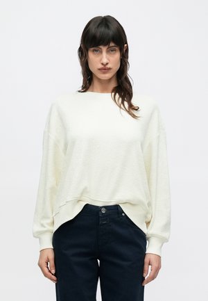 Cremefarbener, strukturierter Pullover mit lässiger Passform, ausgestattet mit geripptem Saum und Bündchen, kombiniert mit dunkelblauen Hosen.