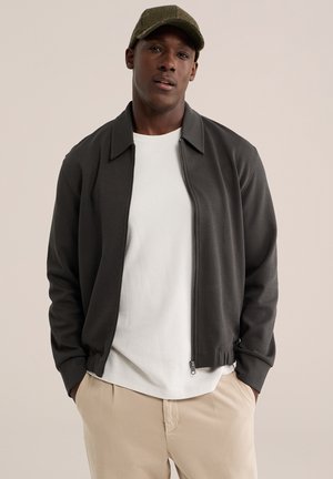 Mann trägt eine olivgrüne Kappe, eine dunkelgraue Zip-up-Jacke über einem weißen Hemd und beige Hose, steht mit den Händen in den Taschen.