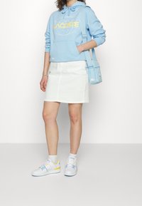 Sweat à capuche bleu clair avec logo "LACOSTE", jupe en jean blanche, sac bleu clair et baskets blanches avec des accents bleus et jaunes.