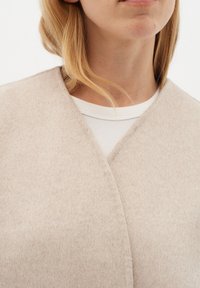 Šviesiai smėlinis vilnos kardiganas su apvyniojimo dizainu, minkšta tekstūra ir V formos iškirpte, dėvimas virš baltos juostuotos marškinių.