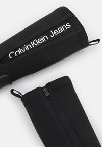 Sort neoprenet accessory med lynlåse, der fremhæves tydeligt. "Calvin Klein Jeans" branding i hvid langs siden. Glat tekstur, minimalistisk design.