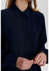 Chemisier bleu marine avec col, manches à poignets plissés et texture de tissu lisse. Présente cinq boutons sur le devant et une coupe classique.