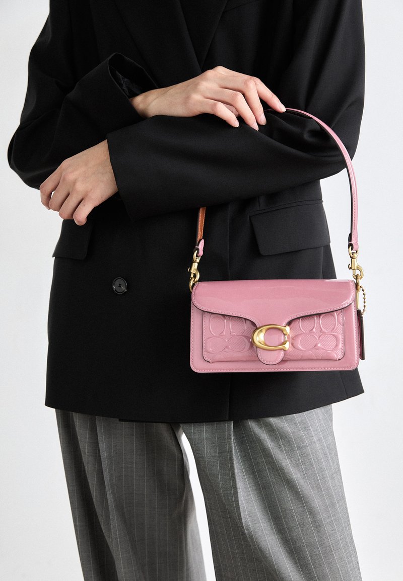 Roosa nahast käekott tekstuuriga tikandi mustriga, kuldne riistvara ja lühike rihm, hoitud musta bleiseri ja triibuliste pükste vastu.