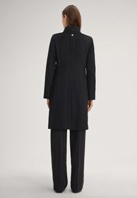 Zwarte lange jas met een gestructureerde kraag, textuurstof en een enkele knop aan de achterkant. Gepaard met zwarte wijde broek.