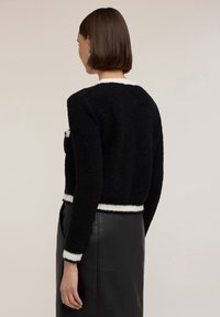 Svart, texturerad tröja med vita randiga mudd och fåll, som visar upp en kortad design. Modellen bär en svart läderskjort.