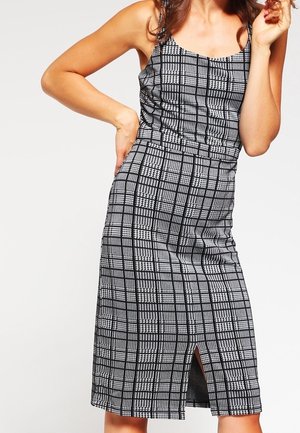 Vrouw draagt een mouwloze zwart-wit geruite midi-jurk met een split aan de voorkant, poseert met één hand op haar heup en de andere bij haar gezicht.