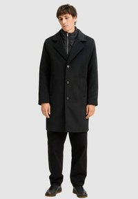 Schwarzer Mantel mit Reverskragen, Knopfleiste und Seitentaschen, getragen über einer schwarzen Zip-Jacke und kombiniert mit schwarzen Hosen und Schuhen.