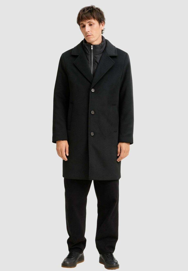 Schwarzer Mantel mit Reverskragen, Knopfleiste und Seitentaschen, getragen über einer schwarzen Zip-Jacke und kombiniert mit schwarzen Hosen und Schuhen.