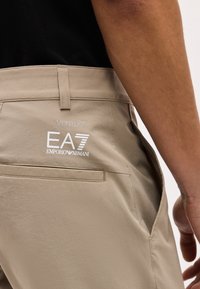 Μπεζ παντελόνι με το λογότυπο "VENTUS EA7 EMPORIO ARMANI" στην πίσω τσέπη, συνδυασμένο με μαύρο πουκάμισο, κοντινό πλάνο στη μέση και το χέρι.