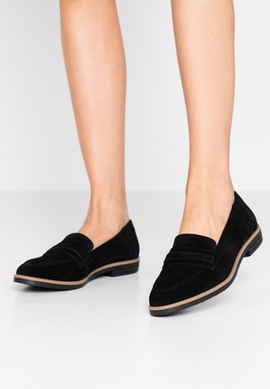 Jambes portant des mocassins en daim noir avec des semelles beiges, debout sur une surface blanche lisse avec un fond clair uni.