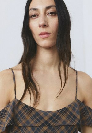 Femme aux longs cheveux foncés portant une robe à bretelles fines à carreaux marron et bleu marine, regardant directement la caméra avec une expression neutre.