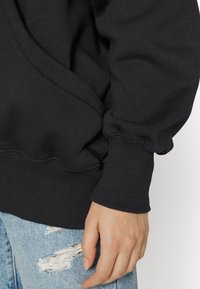 Svart sweatshirt med ribbad mudd, med en texturerad tyg och en sömlös design. En hand visas vilandes på slitna blå jeans.