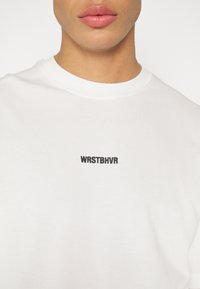 T-shirt en coton blanc avec un col rond et un texte brodé noir "WRSTBHVR" sur le côté gauche de la poitrine. Texture lisse.