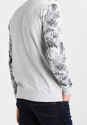 Hellgraues Sweatshirt mit schwarzen und weißen Ärmeln mit Blumenmuster, getragen von einer Person, die von hinten gezeigt wird, kombiniert mit dunkler Jeans.