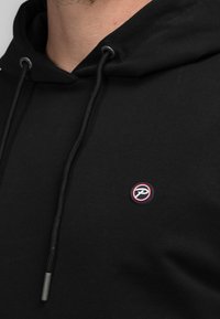 Sudadera negra hecha de tela suave, con cordón ajustable y un pequeño logotipo bordado con acentos en rojo y blanco en el lado izquierdo del pecho.