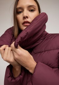 Vínová prošívaná bunda s vysokým límcem, s prošívaným vzorem a měkkou texturou. Model drží límec blízko u těla, což ukazuje detaily rukávu.