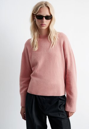 Light pink knit sweater with a ribbed hem and cuffs, featuring a relaxed fit and soft texture. Worn with black trousers.