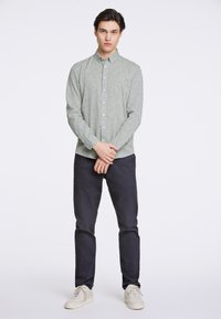 Camisa de botones de manga larga de color verde claro con un patrón texturizado, combinada con pantalones oscuros y zapatillas beiges. El modelo está de pie con las manos entrelazadas.