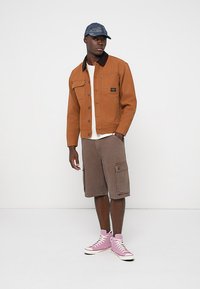 Giacca marrone con colletto nero, camicia bianca, pantaloni cargo marroni e scarpe alte rosa; un cappellino di jeans completa il look.