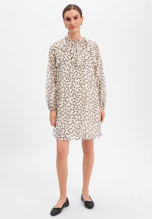 Vrouw draagt een lichte, doorschijnende jurk met zwarte stippen, lange mouwen, gerimpelde halslijn en zwarte platte schoenen, staand op een witte achtergrond.