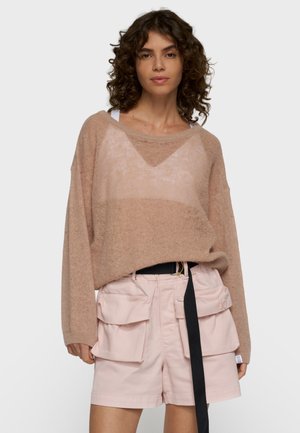 Frau mit lockigem Haar, die einen lockeren beigen Pullover und hellrosa Cargo-Shorts mit großen Taschen und einem schwarzen Gürtel trägt.