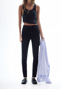 Pantalones vaqueros ajustados negros con una cintura alta, combinados con una camiseta sin mangas oscura. La persona sostiene una camisa de rayas azul claro y lleva mocasines negros.