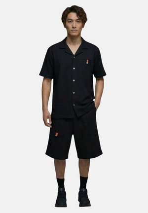Homme portant une chemise noire à manches courtes boutonnée et un short assorti jusqu'aux genoux avec des chaussettes noires et des baskets sur un fond uni.
