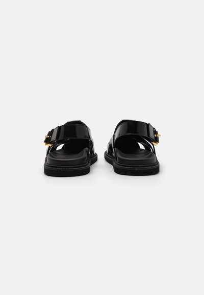 Black patent leather sandals with a low heel, wide strap, and gold buckle detail. Smooth texture with an open back design.