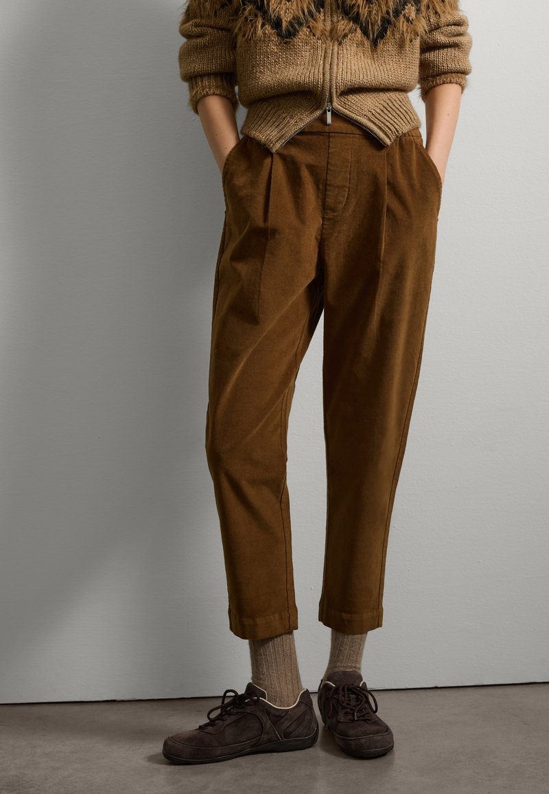 Pantaloni in velluto a coste marroni con pinces, gambe affusolate e risvolti. Abbinati a scarpe in camoscio scuro e calze di lana. Maglione in maglia accogliente sopra.