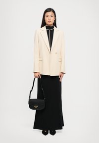 Woman standing against white background wearing cream blazer over black long dress, holding black handbag and wearing black pointed shoes.