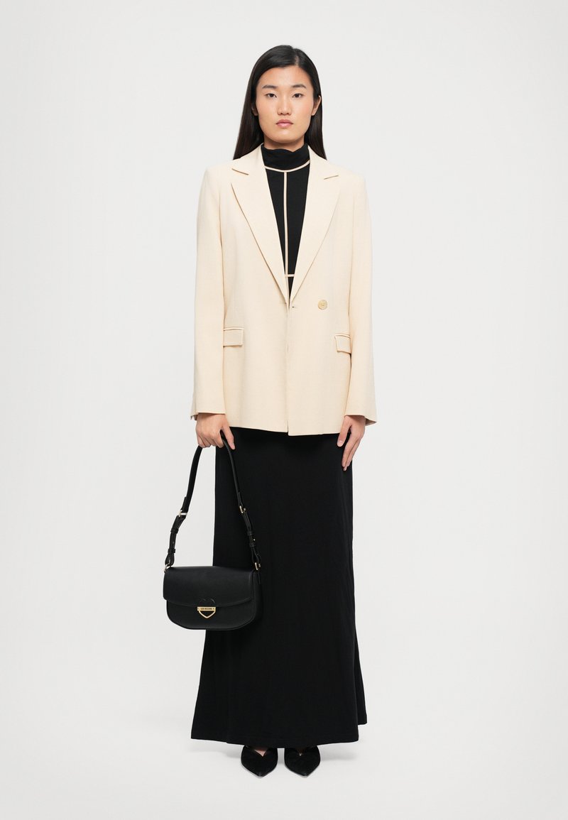 Woman standing against white background wearing cream blazer over black long dress, holding black handbag and wearing black pointed shoes.