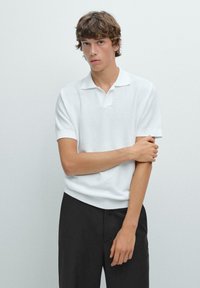 Polo shirt bianco a maniche corte in tessuto strutturato con colletto a piatto, abbinato a pantaloni neri. Il modello si trova di fronte a un muro semplice.