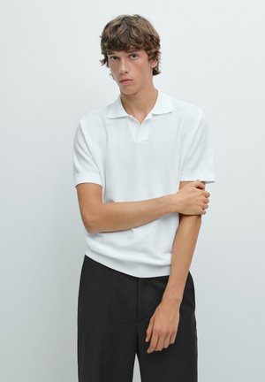 Polo shirt blanc à manches courtes en tissu texturé avec un col plat, associé à un pantalon noir. Le modèle se tient devant un mur uni.