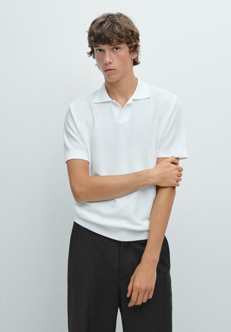 Polo shirt bianco a maniche corte in tessuto strutturato con colletto a piatto, abbinato a pantaloni neri. Il modello si trova di fronte a un muro semplice.
