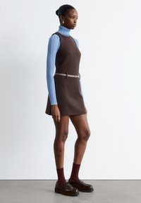 Braunes ärmelloses Kleid mit taillierter Passform, darunter ein hellblauer gerippter Rollkragenpullover, ein silberner Gürtel, kastanienbraune Socken und dunkle Loafers.