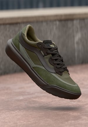Zielono-ciemnobrązowy sneaker Vans ze sznurowadłami, teksturowaną podeszwą i metką Ultrrange, pokazany w powietrzu na różowawym betonowym podłożu.