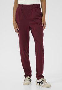 Pantalons ajustés bordeaux avec une texture lisse, présentant une taille haute et des plis. Portés avec des baskets blanches et noires.