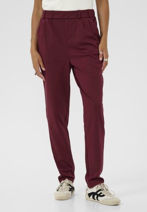 Pantalons ajustés bordeaux avec une texture lisse, présentant une taille haute et des plis. Portés avec des baskets blanches et noires.