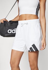 Vita atletiska shorts med elastisk midja och dragsko, med ett svart diagonalt randmönster längs fållen. Svart väska med logotyp.