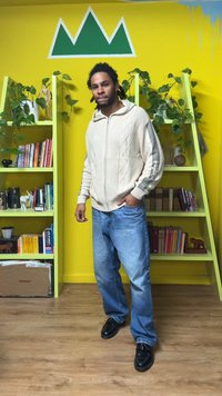 Pull beige à fermeture éclair en maille torsadée, jean bleu et chaussures noires. Pièce aux murs jaunes, étagères vertes et plantes.