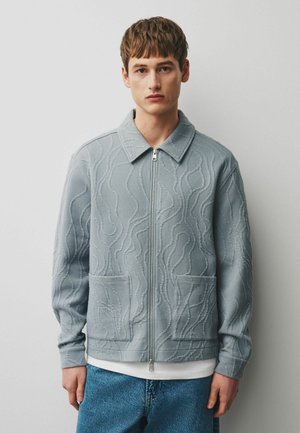 Young man wearing a light gray zip-up jacket with textured abstract patterns and front pockets, paired with blue jeans and a white undershirt.