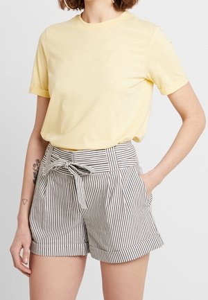 Kvinde iført en bleg gul kortærmet t-shirt og højtaljede shorts i sort-hvide striber med et stofbælte bundet foran.