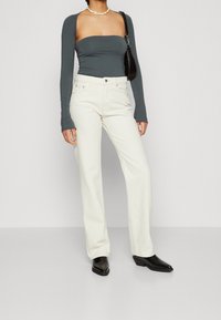 Mujer vestida con una blusa larga asimétrica en gris oscuro, jeans blancos de cintura alta, botas puntiagudas negras, collar de conchas y bolso negro de hombro.