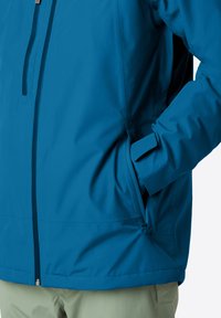 Mano in una tasca parzialmente chiusa di una giacca blu con zip frontale, indossata sopra pantaloni verde chiaro.
