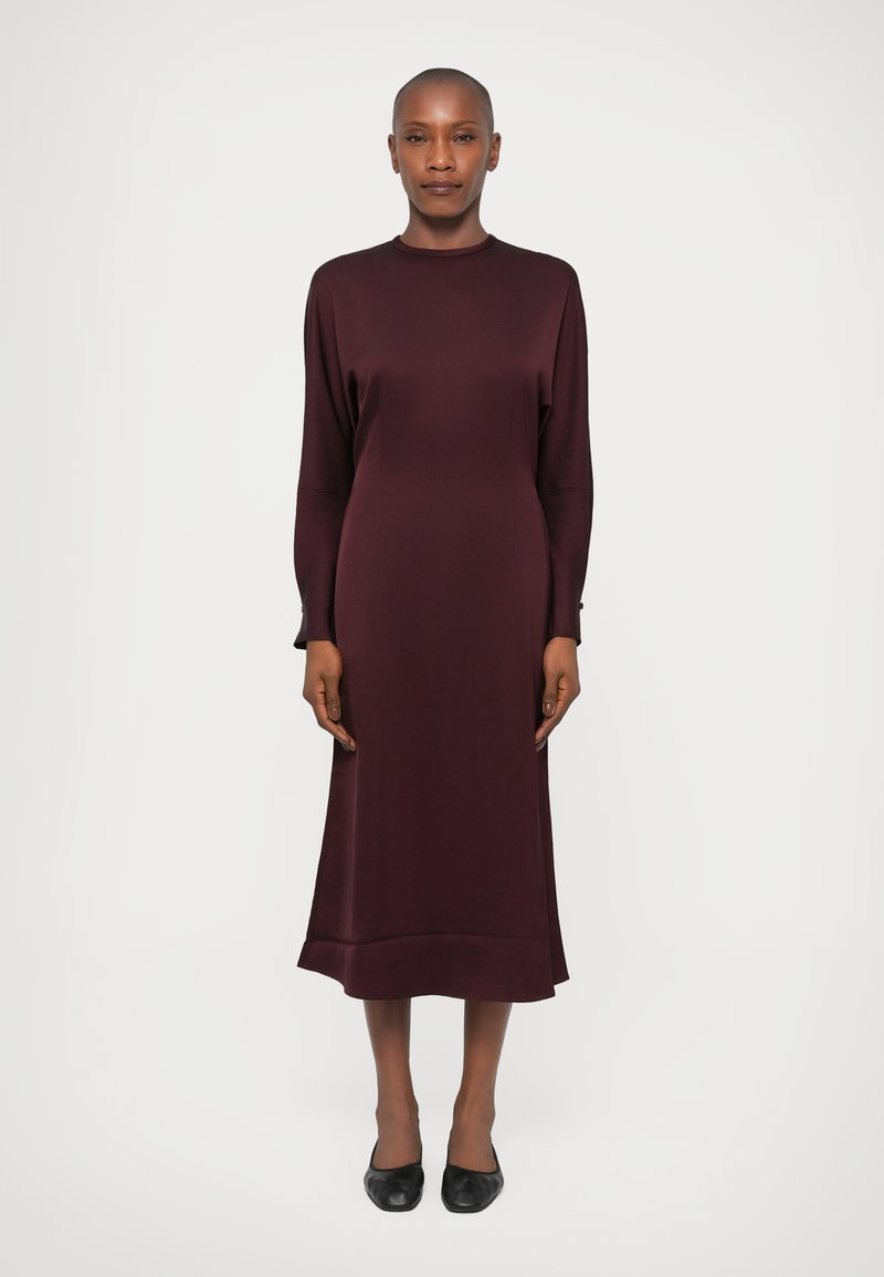 Rochie lungă de culoare burgundă, cu maneci lungi, de tip A, cu guler rotund și țesătură netedă. Purtați cu pantofi plat de culoare neagră, stând pe un fundal neutru.