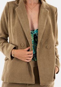 Blazer in corduroy beige con due bottoni, revers a punta e finitura testurizzata, indossato sopra un top colorato a motivo.
