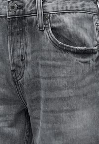 Gros plan sur un jean en denim gris montrant la poche avant, la boucle de ceinture et un bouton en métal avec des coutures visibles et la texture du tissu.