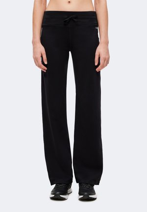 Woman wearing black wide-leg sweatpants with drawstring waist and black athletic shoes, standing against plain background.