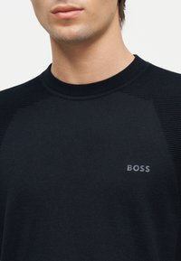 Νέος άνδρας που φοράει μαύρο πλεκτό πουλόβερ με ριμπ ώμους και το λογότυπο "BOSS" στο στήθος.