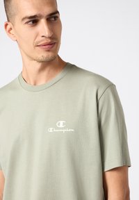 Camiseta de manga corta en verde salvia claro, hecha de algodón suave y con cuello redondo. Presenta un pequeño logo blanco de Champion en el pecho.